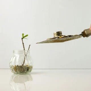 A hand holding a knife cuts a coin from a jar containing a small plant, symbolizing financial growth and investment.